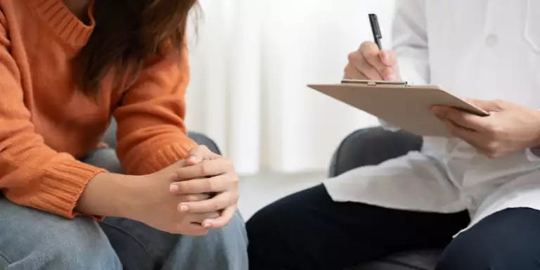 A woman in an orange sweater is sitting on a couch, holding a pen in her hand. She is also holding a clipboard in her other hand. A doctor in a white coat is seated next to her, also holding a clipboard. The doctor is looking at the clipboard, possibly discussing something. The background is a simple white wall.