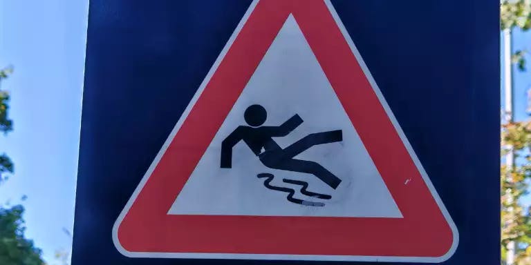 A pedestrian crossing sign with a red border and a white background, featuring a silhouette of a person falling over. The sign is mounted on a blue pole and is set against a backdrop of trees and a clear blue sky.