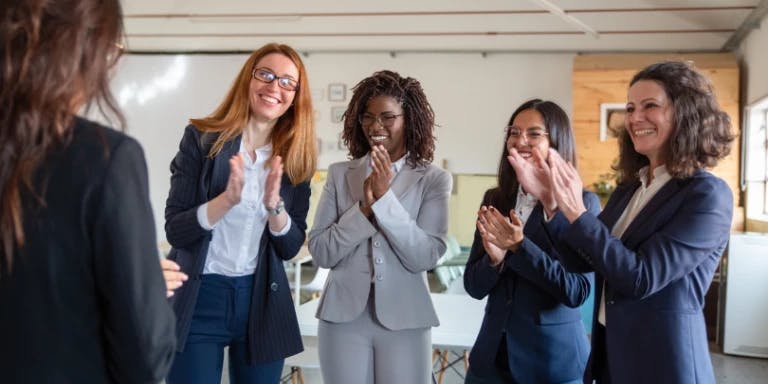 Four women are gathered in a room with a white wall and a window. They are all dressed in business attire, including blazers and suits. The woman on the left is clapping her hands, while the woman in the middle is smiling and looking at the camera. The woman on the right is also clapping her hands, and the woman on the far left is looking at the camera. They are all standing in front of a whiteboard. Four women are gathered in a room with a white wall and a window. They are all dressed in business attire, including blazers and suits. The woman on the left is clapping her hands, while the woman in the middle is smiling and looking at the camera. The woman on the right is also clapping her hands, and the woman on the far left is looking at the camera. They are all standing in front of a whiteboard.