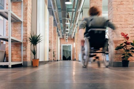 A person in a wheelchair is moving towards the right side of a hallway. The hallway has brick walls and a gray floor. There are potted plants on the left side of the hallway. A doorway is visible on the right side of the hallway. The person in the wheelchair is blurred, indicating motion.