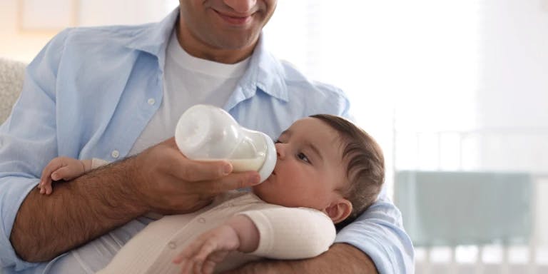 A man in a blue shirt is holding a baby in his arms, feeding the baby with a pacifier. The baby is wearing a white onesie and is sitting on a white couch. The background features a window with white curtains and a white rail. A man in a blue shirt is holding a baby in his arms, feeding the baby with a pacifier. The baby is wearing a white onesie and is sitting on a white couch. The background features a window with white curtains and a white rail.