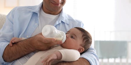 A man in a blue shirt is holding a baby in his arms, feeding the baby with a pacifier. The baby is wearing a white onesie and is sitting on a white couch. The background features a window with white curtains and a white rail.