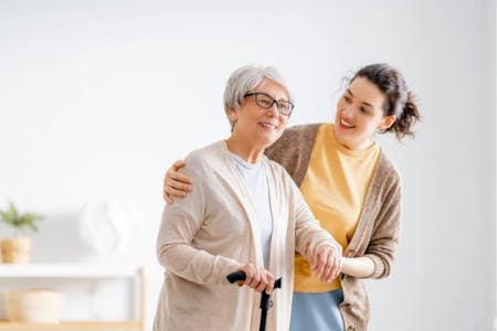 Two elderly women are in a room with a white wall and a wooden floor. The woman on the left is wearing a beige cardigan and glasses, while the woman on the right is wearing a yellow sweater and has a cane in her hand. They both appear to be laughing and smiling, suggesting a positive interaction. The woman on the right is holding the woman on the left's hand, indicating a close relationship. The woman on the left is also wearing a scarf, adding to the cozy atmosphere of