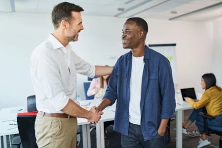 In the image, there are two men in an office setting. The man on the left is dressed in a white shirt and khaki pants, while the man on the right is wearing a blue shirt and khaki pants. They are shaking hands, indicating a positive interaction. The office has white walls and a desk with a computer monitor and a whiteboard. In the background, there are other people seated at desks, suggesting a collaborative work environment. The man on the left is smiling, indicating a