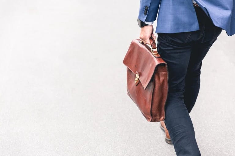 A man in a blue suit is walking on a gray sidewalk, carrying a brown leather briefcase with a gold zipper. The man's posture suggests he is moving forward, possibly heading to or from a destination. The background is blurred, making it difficult to discern specific details. The man's attire and the briefcase indicate a professional setting.