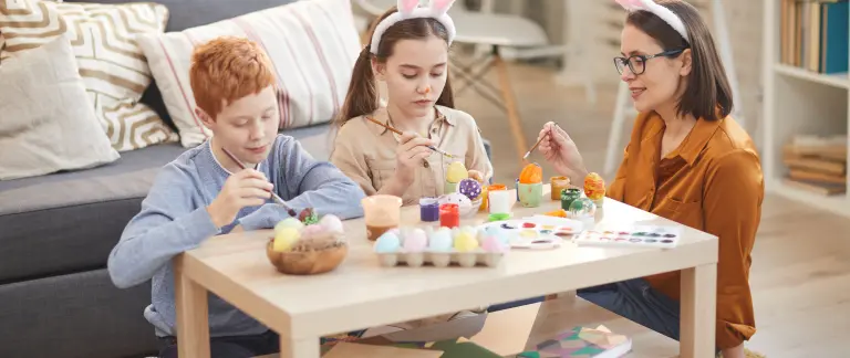 A family of three is enjoying a creative activity in a cozy living room. The mother, wearing glasses, is seated on a gray sofa, engrossed in a painting. The father, with red hair, is seated on the coffee table, holding a paintbrush. The daughter, with red hair, is seated on the floor, also holding a paintbrush. The coffee table is laden with various art supplies, including eggs, paintbrushes, and paint. The room is filled with soft lighting
