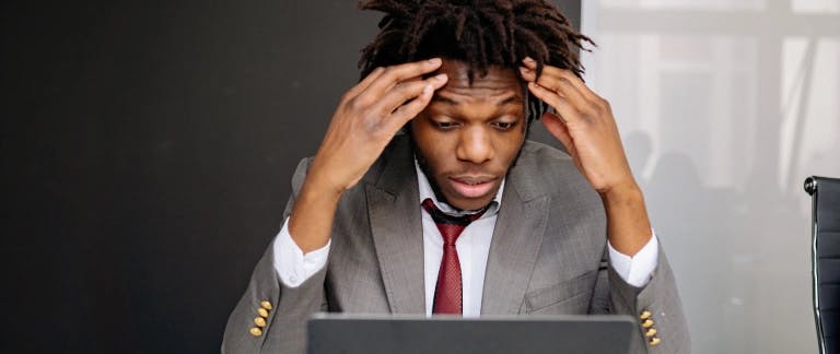 A worried-looking man wearing a suit, sitting in front of a laptop with his hands on his forehead