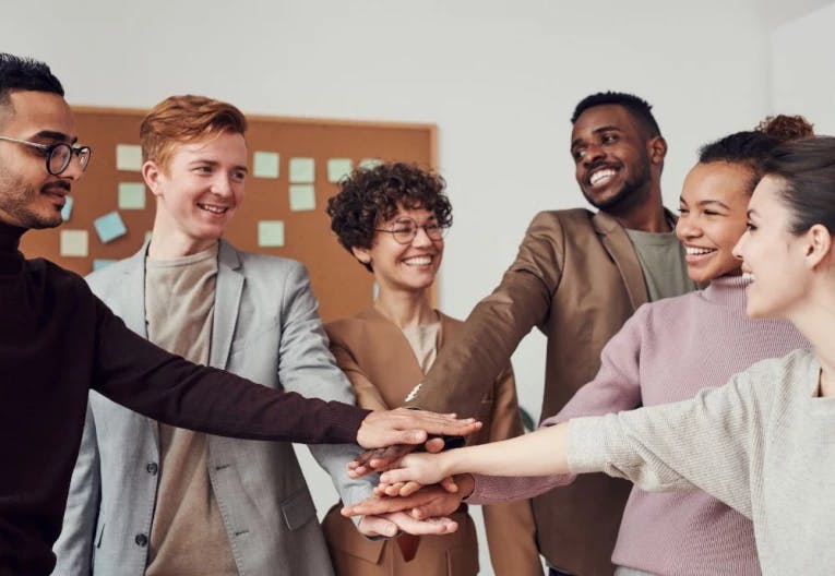A group of six individuals, dressed in business attire, are gathered in a room with a bulletin board in the background. They are holding hands and smiling, indicating a positive interaction. The room has a white wall and a wooden floor, and there are sticky notes scattered around the room. The overall atmosphere suggests a team-building event or a celebration.