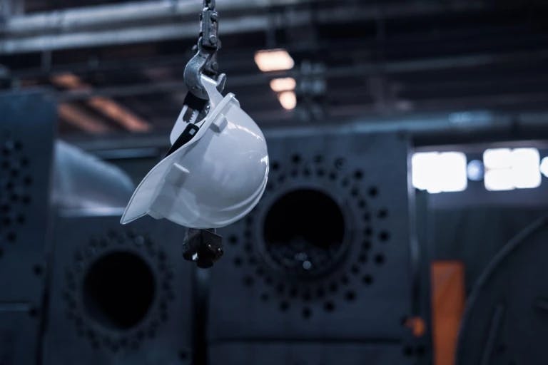 A white hard hat is suspended from a hook in a factory setting. The hard hat is attached to a metal chain, which is suspended from a hook in the foreground. The background features large industrial machinery, including pipes and a large cylindrical object. The machinery is illuminated by bright lights, and there's a sign with the letter "K" visible in the background. The image captures a moment in a factory, with the hard hat as the central focus.
