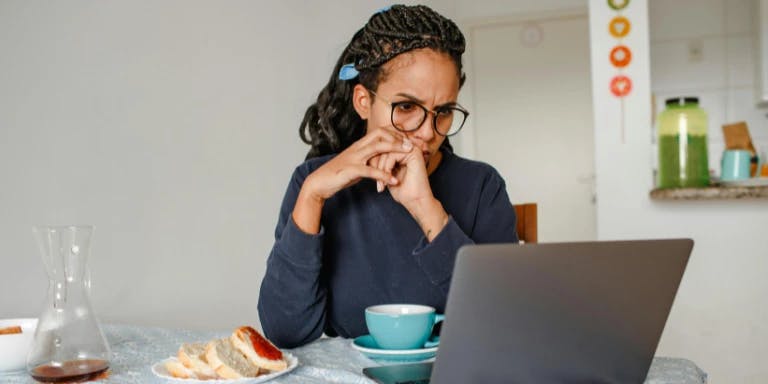 A woman with glasses is seated at a table, engrossed in her laptop. She is wearing a blue sweater and has her hand on her chin. The table has a plate of bread and a glass of water. The background features a white wall with a shelf holding various items.