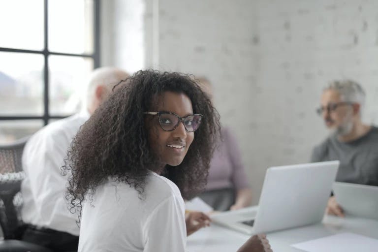 A woman with curly hair and glasses is seated at a table in a room with a white brick wall. She is smiling and appears to be engaged in a meeting or discussion. Behind her, two men are seated at the table, both wearing gray shirts. One of them is holding a laptop, suggesting they might be working together. The room has a window, allowing natural light to enter. The woman's position and the men's actions suggest a collaborative work environment.