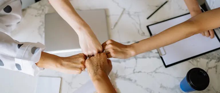 Two people are holding hands in a professional setting, possibly in a meeting or collaboration. The background features a marble table with a laptop and a cup, suggesting a workspace or office environment.