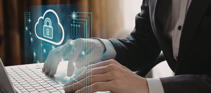 A person in a suit is typing on a laptop computer, which is placed on a desk. The laptop screen displays a blue cloud with a lock symbol on it. The person's hands are interacting with the keyboard, and the background is a blurred image of a computer network.