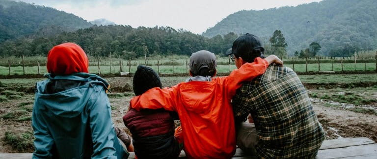 A photo of four people sitting on a bench with their backs to the camera and looking at forest-covered hills