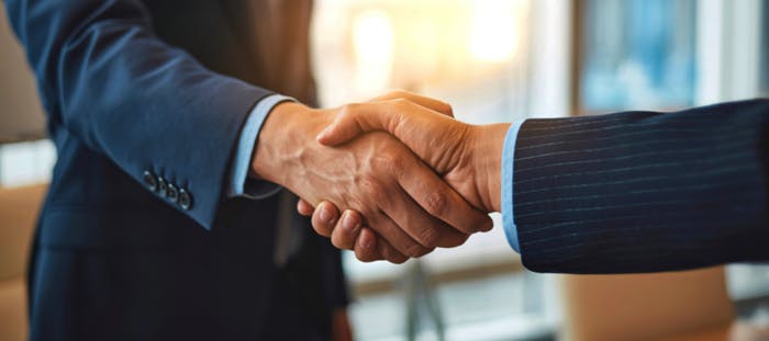 Two individuals in business suits are shaking hands in a professional setting. The background is blurred, but it appears to be an office or a conference room with a window. The image captures a moment of agreement or collaboration between the two individuals.