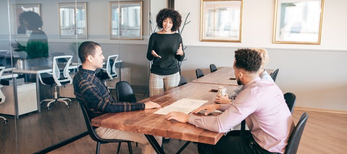 A woman in a black shirt is standing at a long wooden conference table, holding a pen and a piece of paper. She is surrounded by two men, one with curly hair and the other with short hair. The room has windows and a white wall with framed pictures.
