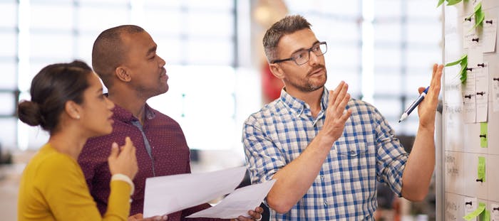 Three individuals are gathered around a whiteboard in an office setting. The person on the left is wearing a blue and white plaid shirt, holding a piece of paper and appears to be in the middle of a discussion. The person in the middle is wearing a yellow shirt and is holding a piece of paper as well. The person on the right is wearing a red shirt and is also holding a piece of paper. They are all looking at the whiteboard, which is covered with green sticky notes.