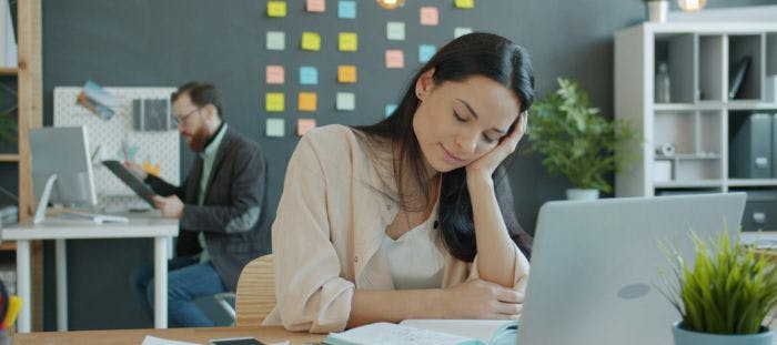 A woman is sitting at a desk in an office, working on a laptop. She is resting her head on her hand and appears to be in a state of deep thought. The desk is cluttered with papers and a plant. In the background, there is a man standing behind the desk, possibly working or waiting for his turn to use the laptop. The office has a wall with colorful sticky notes, adding a touch of personality to the workspace.