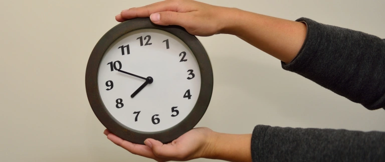 A hand is holding a round clock with a black frame, displaying the time as 10:10. The hands are positioned at 10 and 3, indicating the time. The background is a plain white wall.