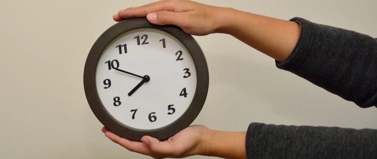 A hand is holding a round clock with a black frame, displaying the time as 10:10. The hands are positioned at 10 and 3, indicating the time. The background is a plain white wall.