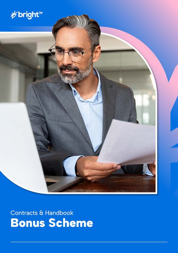 A man with a beard and glasses is seated at a desk, holding a piece of paper. He is dressed in a gray suit and blue shirt, and is wearing glasses. The background features a blue and white gradient.