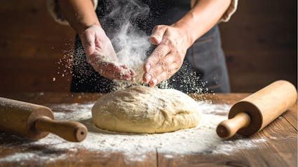Wild Hearth Bakery A person is preparing a pizza dough on a wooden table. They are using flour to sprinkle it with white powder, and rolling it out on a rolling pin. There are two rolling pins nearby. The person's hands are holding the dough, and they are wearing an apron.