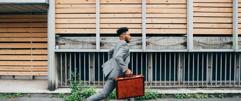 A man in a gray suit is walking on a sidewalk, carrying a brown briefcase. He is moving towards the right side of the image. The background features a wooden wall with vertical slats and a metal fence. The man appears to be in a professional setting, possibly in a business or office environment.