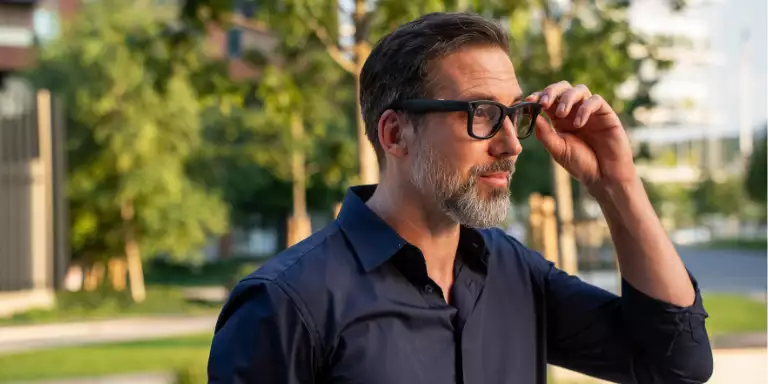 A man stood in a green space wearing meta glasses using the touch features on the left-hand side frame.