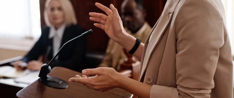 A photograph of a person speaking into a microphone with two people sitting behind her taking notes