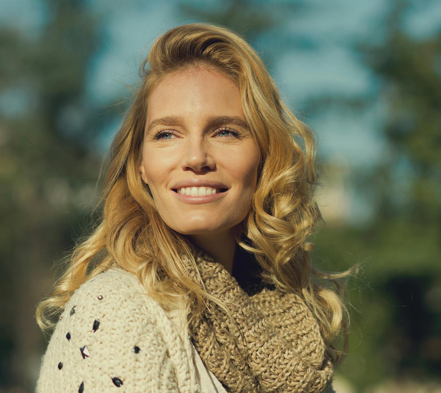 Blond woman wearing a scarf with trees in the background.