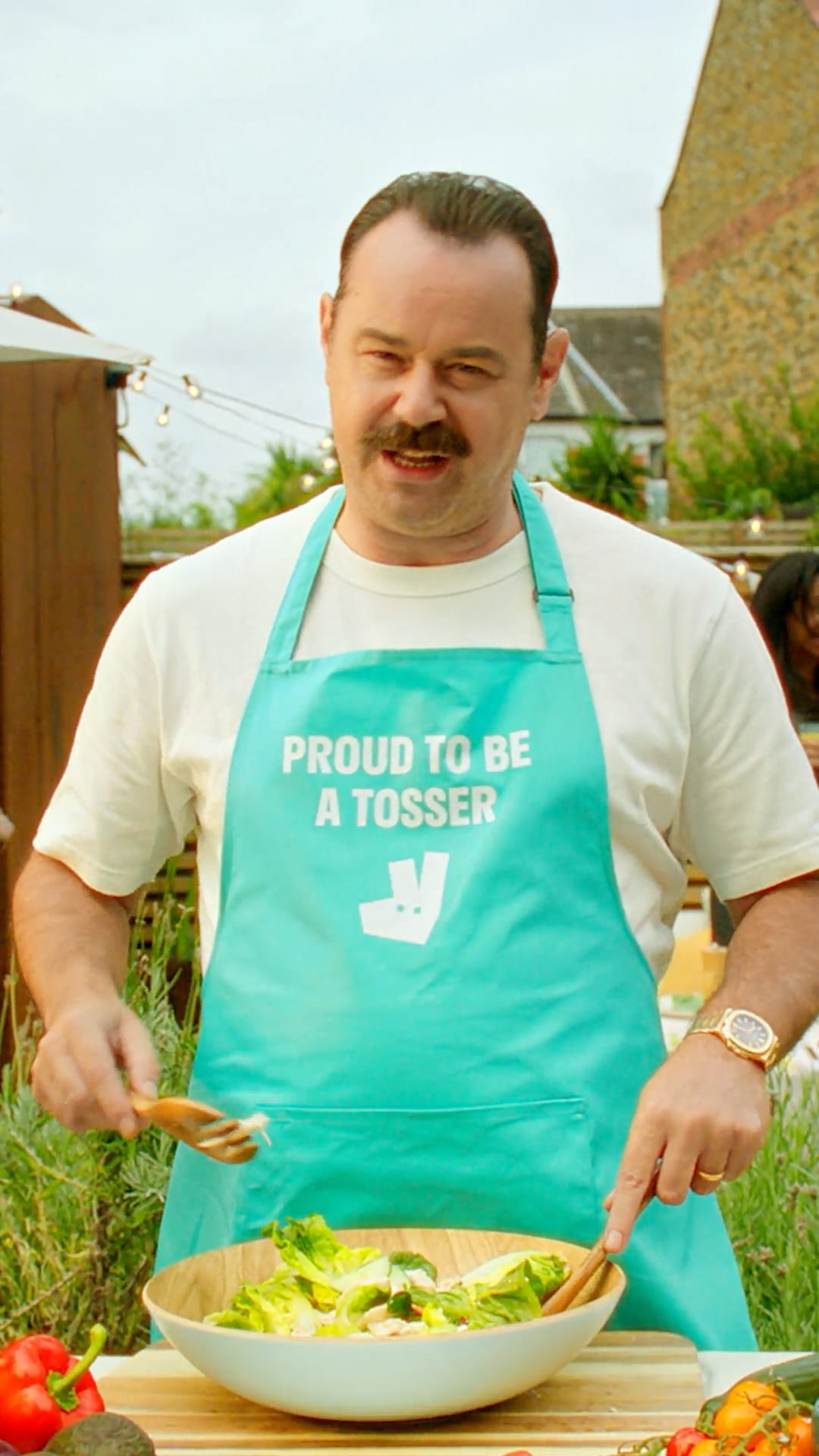 Danny Dyer in a Proud To Be A Tosser apron tossing a salad for Deliveroo