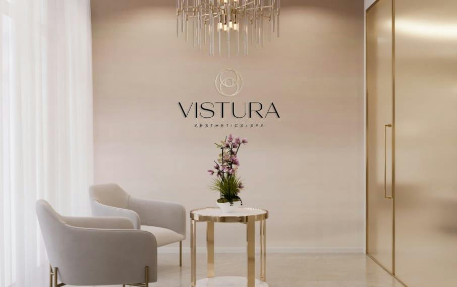 Vistura Aesthetics + Spa reception with beige armchairs, gold side table, orchid, chandelier and logo on wall