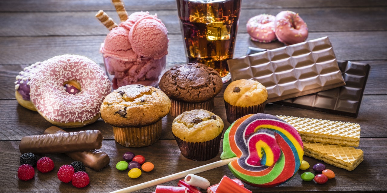 Assortment of sugary foods like candies, gummy candies, donuts, soda, chocolate, lollipop, wafers and cupcakes.