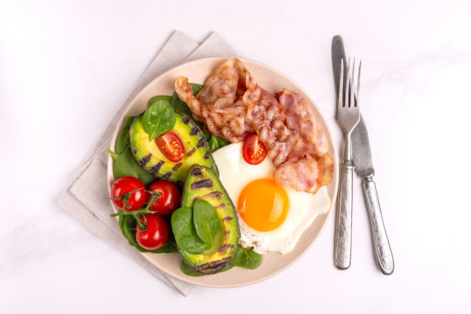 Keto breakfast with grilled bacon and avocado, fried eggs, spinach and cherry tomatoes