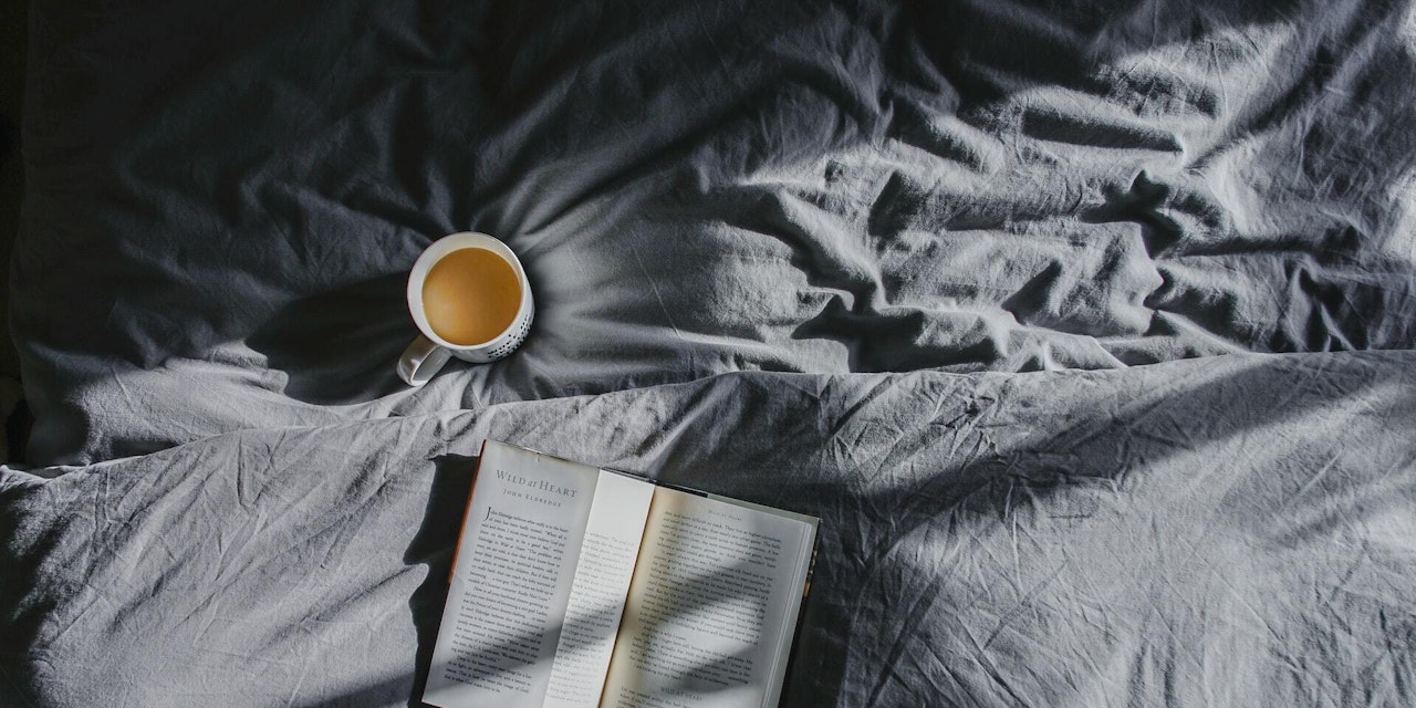 A cup of coffee and a book on a bed strewn with sunlight