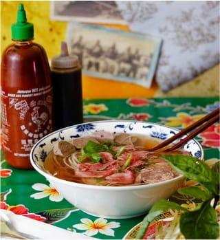 Soupe pho au bœuf saignant sur une nappe fleurie, à côté d’une bouteille de sriracha