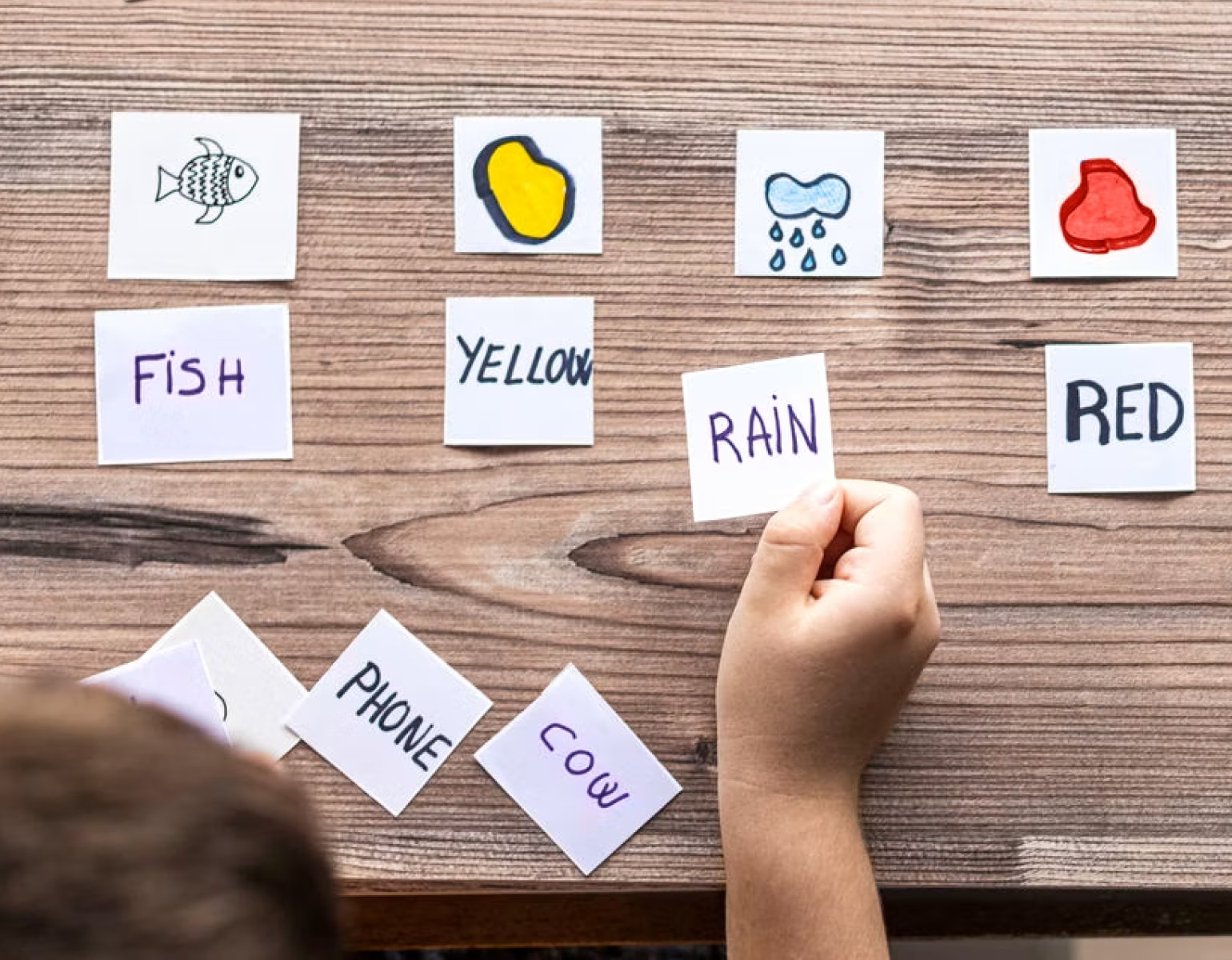 Word and picture cards on a wooden table, with a child's hand