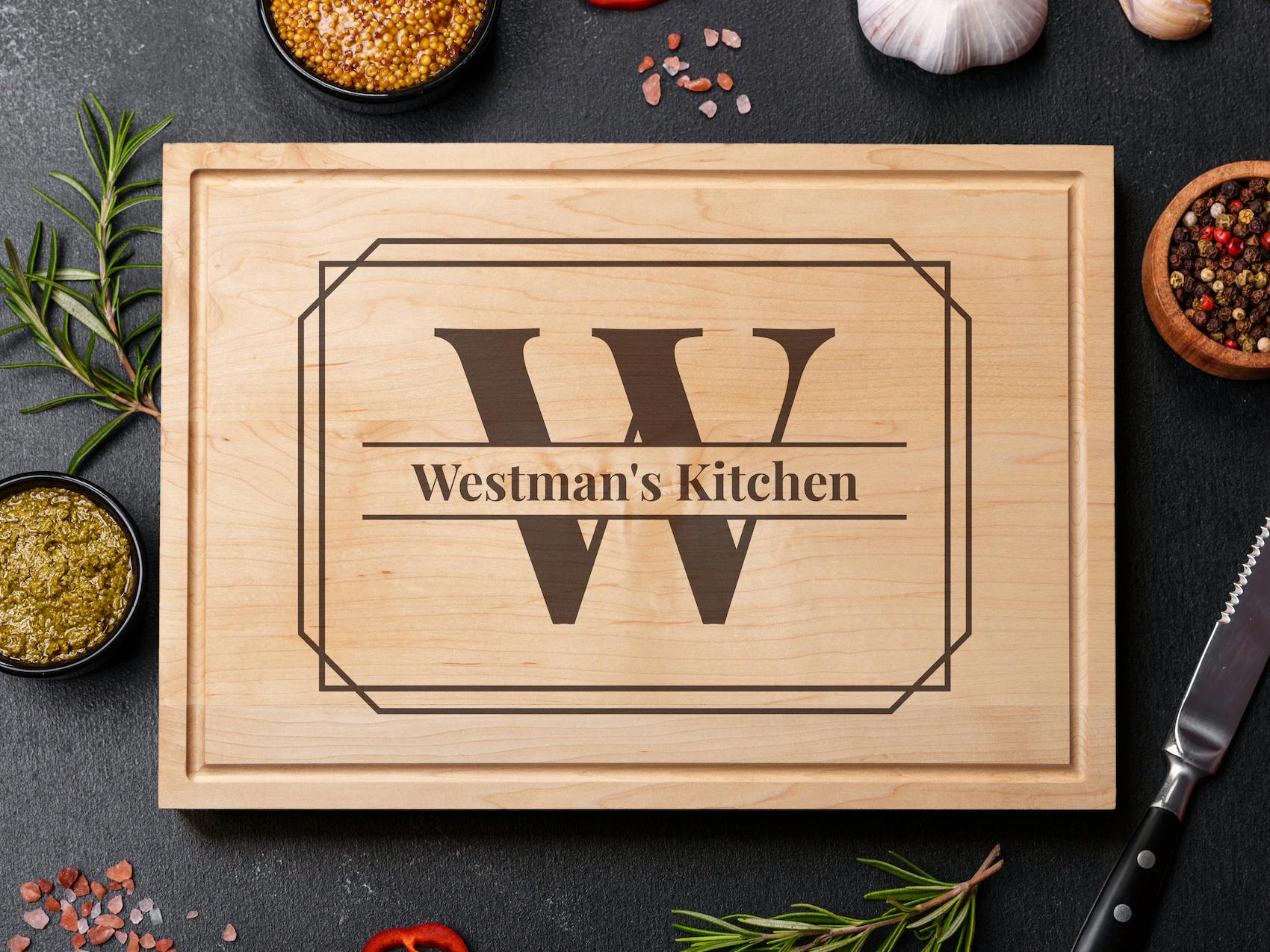 Personligt skærebræt i lyst træ med et stort graveret 'W' og teksten 'Westman's Kitchen' omgivet af krydderier og køkkenredskaber.