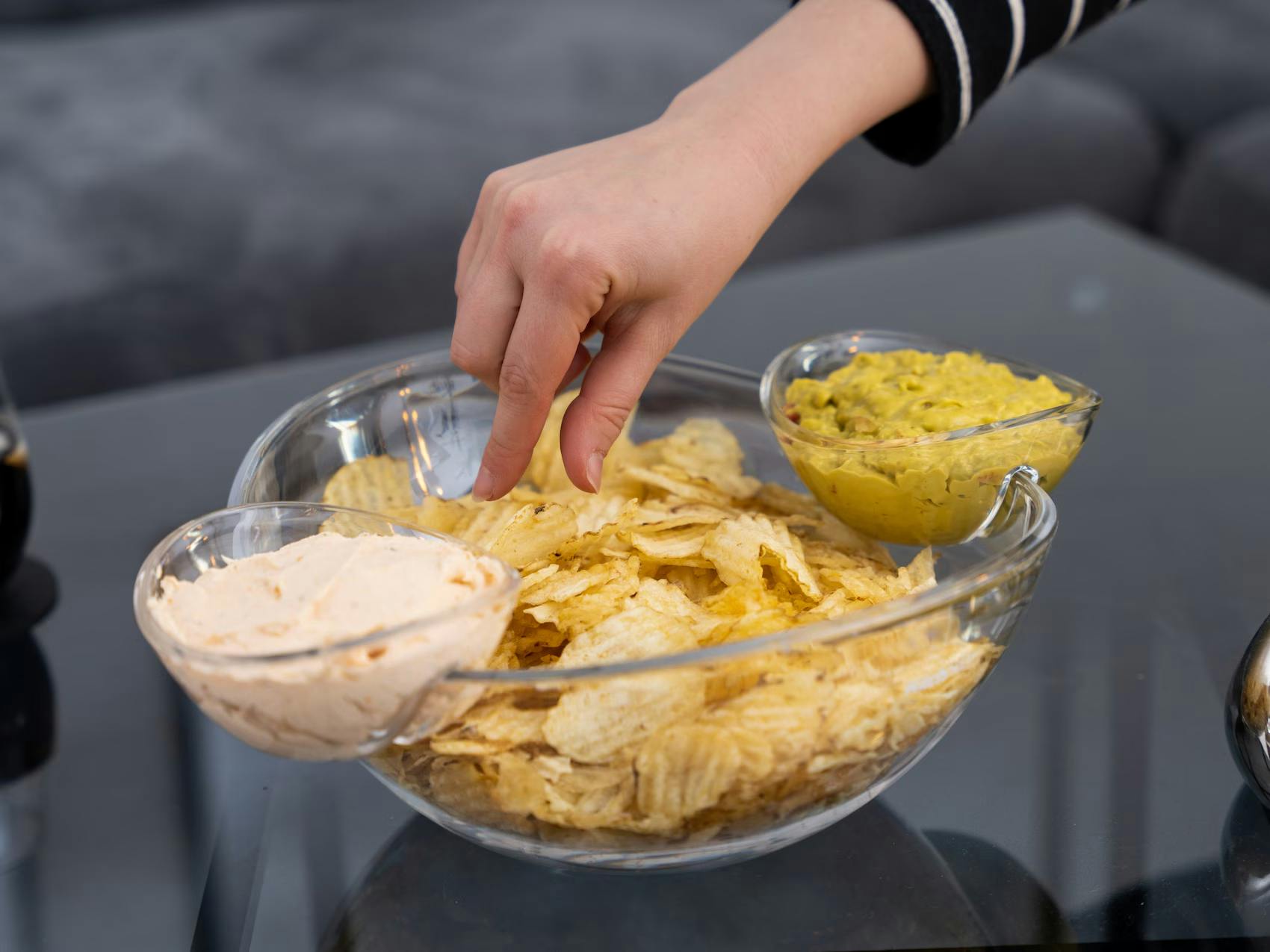KitchPro stor chipsskål i glas med två smarta, avtagbara dippskålar på sidorna. En hand doppar ett chips under fotbollsfesten.