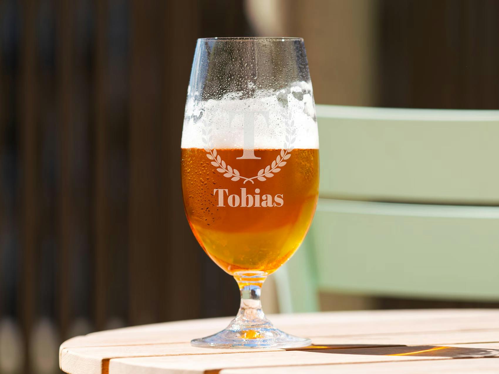 Ett elegant, graverat ölglas med namnet Tobias fyllt med öl, stående på ett bord utomhus i solsken.