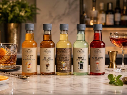 Fem flasker med cocktailmix i forskellige smagsvarianter stillet op på en marmorbordplade, omgivet af elegante drinkglas og barudstyr.