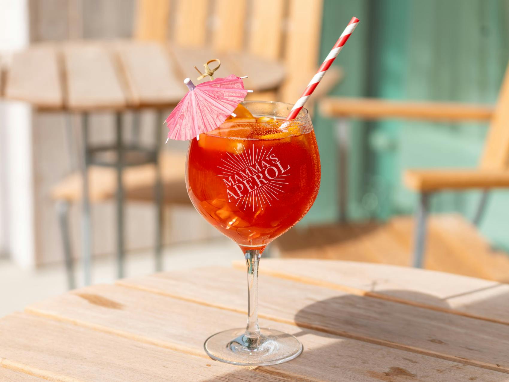 Et stort krystalglas med graveringen "Mamma's Aperol" fyldt med en orange Aperol Spritz, is, et rød- og hvidstribet sugerør og en lille lyserød drinkparasol på et lyst træbord.