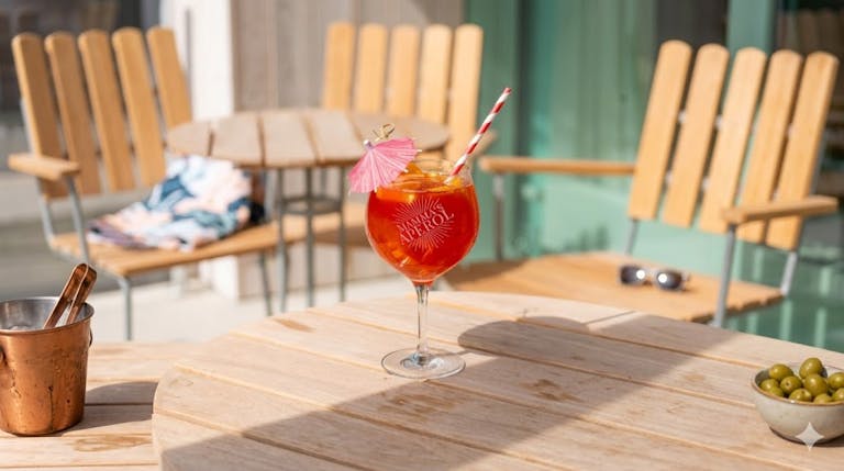 Et stort krystalglas med graveringen "Mamma's Aperol" fyldt med en orange Aperol Spritz, is, et rød- og hvidstribet sugerør og en lille lyserød drinkparasol på et lyst træbord.