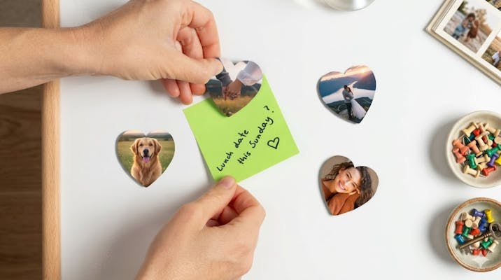 Eine horizontale Ansicht eines weißen Schreibtisches, auf dem zwei Hände in der Mitte eine grüne Haftnotiz mit der Aufschrift „Lunch date this Sunday?“ halten. Um die Notiz herum befinden sich vier herzförmige Fotomagnete mit persönlichen Motiven sowie Schreibtischzubehör wie ein Holz-Organizer, Bücher und ein Fotoalbum.