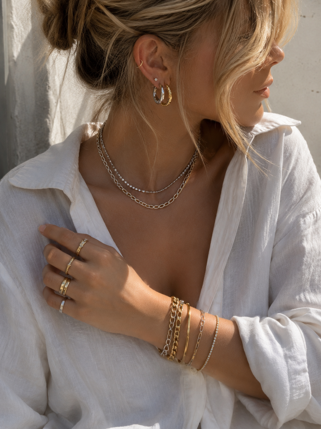 Woman in a white shirt wearing layered silver and gold necklaces with stacked rings and bracelets