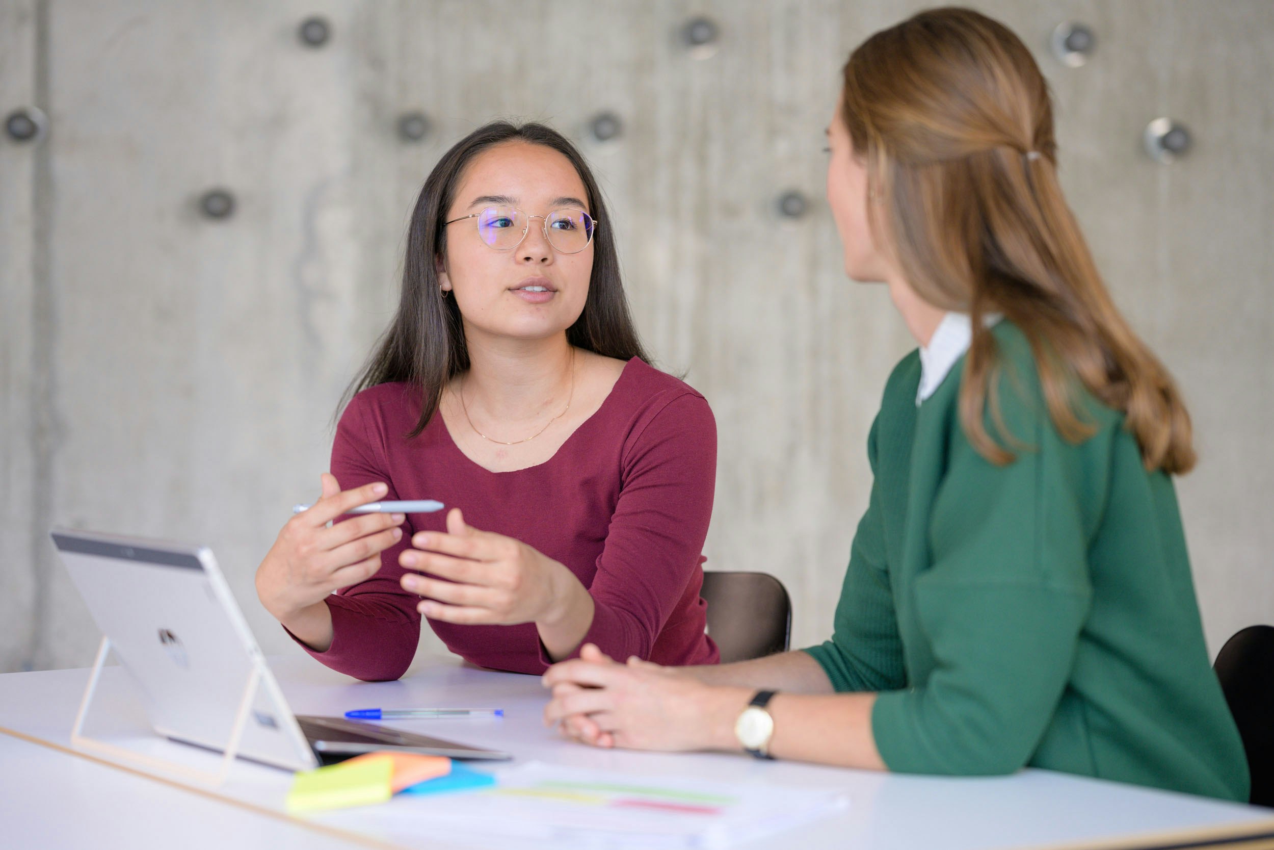 Eine Lernende des Bildungsgangs Bank für IT (BankFIT) tauscht sich mit einer begleitenden Person an einem Tisch mit diversen Arbeitsmaterialien wie zum Beispiel Laptop, Post-its und Stift aus.