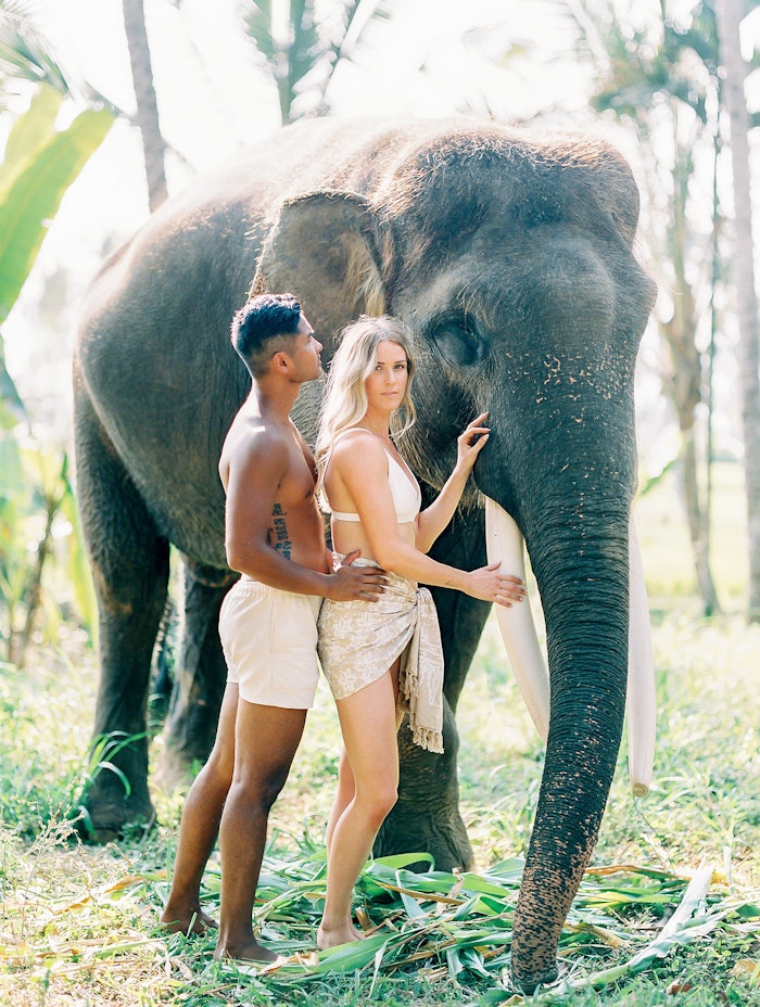 Engaged couple standing close together beside an elephant in Bali, captured in glowing natural light with a soft editorial feel