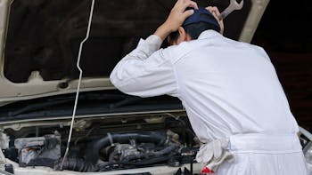 Side view of frustrated stressed young mechanic man in white uniform touching his head with hands against car in open hood at the repair garage. 