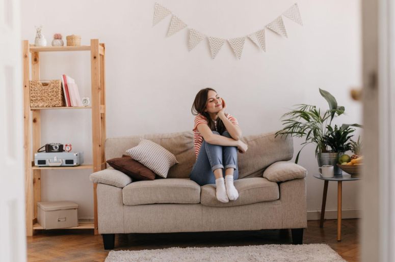 Pessoa sentada em sofá para sala pequena, com almofadas decorativas, estante de madeira, mesa lateral com plantas e decoração aconchegante em ambiente compacto.
