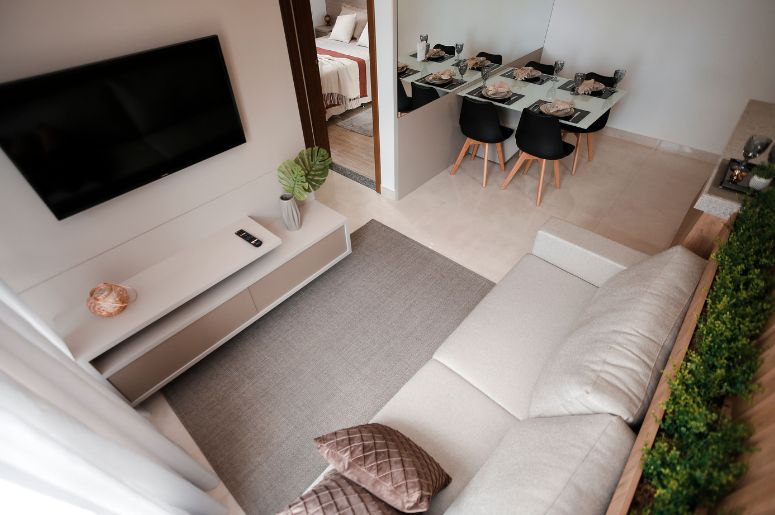 Rack para sala pequena com TV suspensa na parede, sofá claro em L, tapete neutro, mesa de jantar ao fundo e decoração compacta